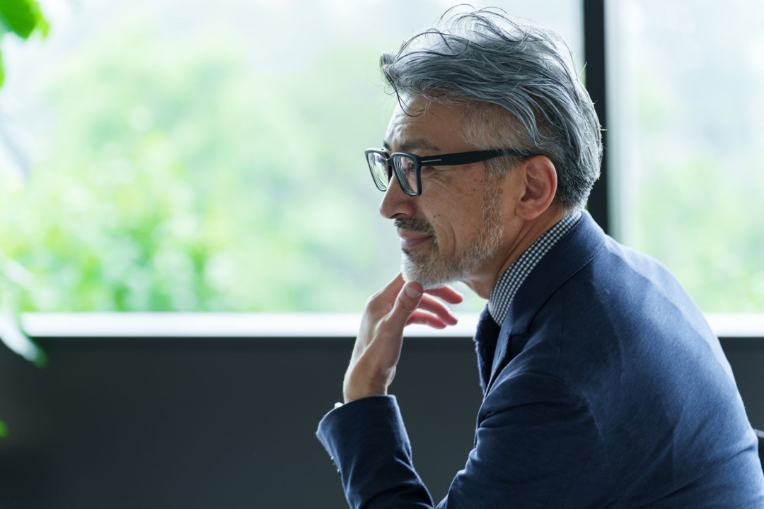 Side profile of a thoughtful man with glasses and gray hair, sitting in a bright room with a window and greenery in the background