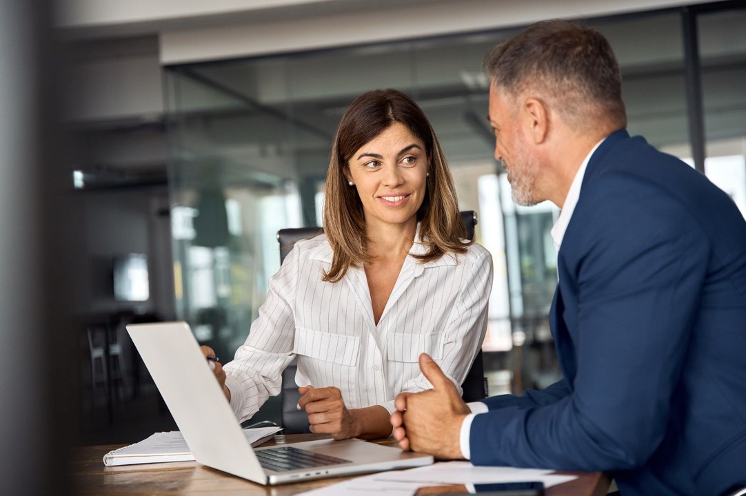 Two colleagues in a modern office discussing work, with a woman smiling and using a laptop while talking to a man in a suit