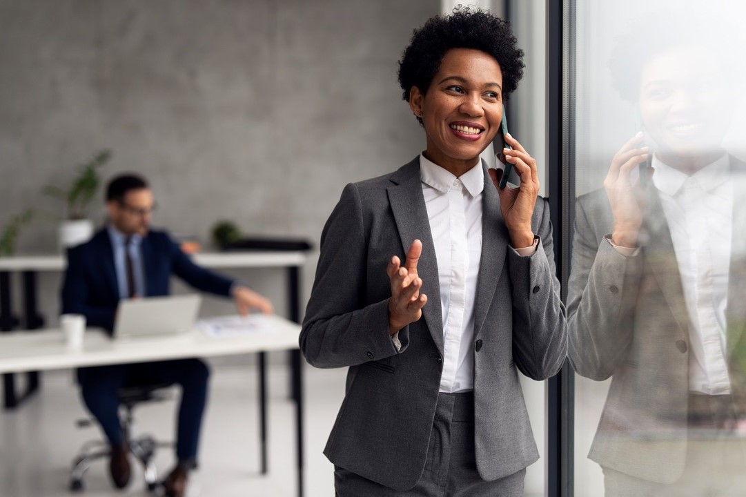 A businesswoman in a gray suit smiling while talking on the phone by a window, with a colleague working at a desk in the background