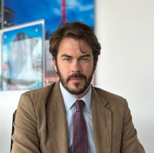 Person in a suit sitting in an office with framed pictures in the background.