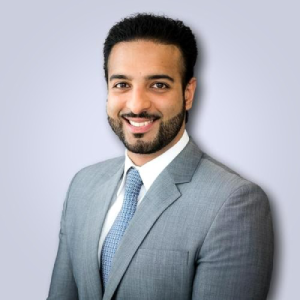 Person smiling in a light grey suit with a blue tie, against a light grey background.