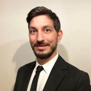 Person in a suit with a polka dot tie smiling in front of a plain background.