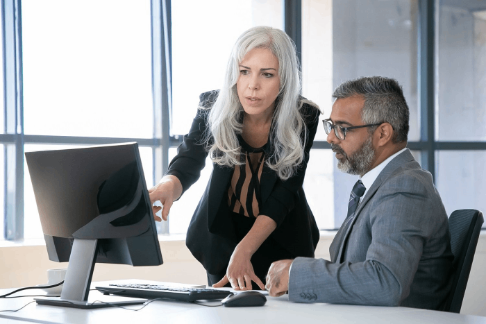 Professional woman with gray hair pointing at a computer screen while discussing with a seated man in a suit.