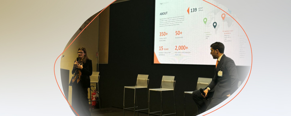 Two people participating in a presentation, with one speaking on stage and a large slide displayed behind them.