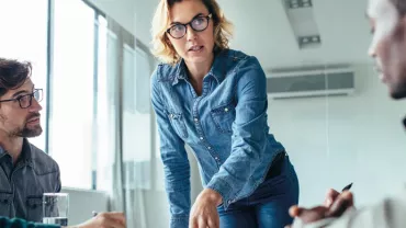 Person in denim speaking at a meeting, with others listening attentively around a table.