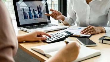 Two people discussing charts and notes at a desk with a laptop.