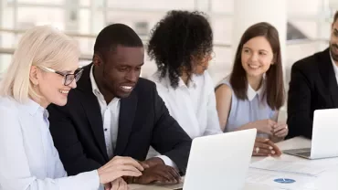 A diverse group of professionals working together on laptops at a bright, modern office table.