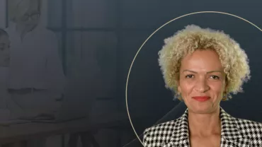  A professional portrait of a woman with curly blonde hair, wearing a patterned blazer, overlaid on a dark background with subtle graphic lines and the faint image of two women working on a laptop in the background.